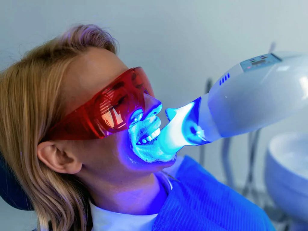 A patient wearing protective red goggles receiving a blue light teeth whitening treatment in a dental office.