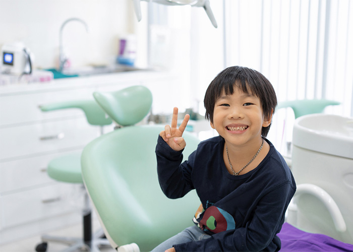 A cheerful young boy sitting in a dental office, flashing a peace sign with a bright smile, showing his missing front teeth. The dental clinic has a modern, clean background with dental chairs and equipment.