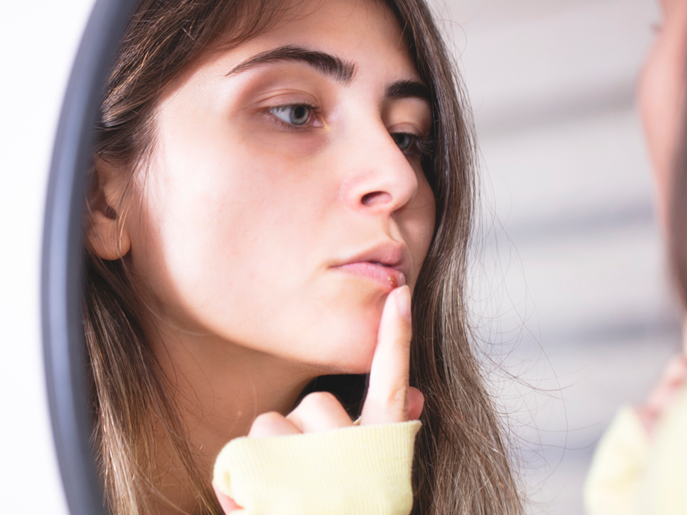 A young woman examining her face in a mirror, touching her mouth gently, checking a cold sore.