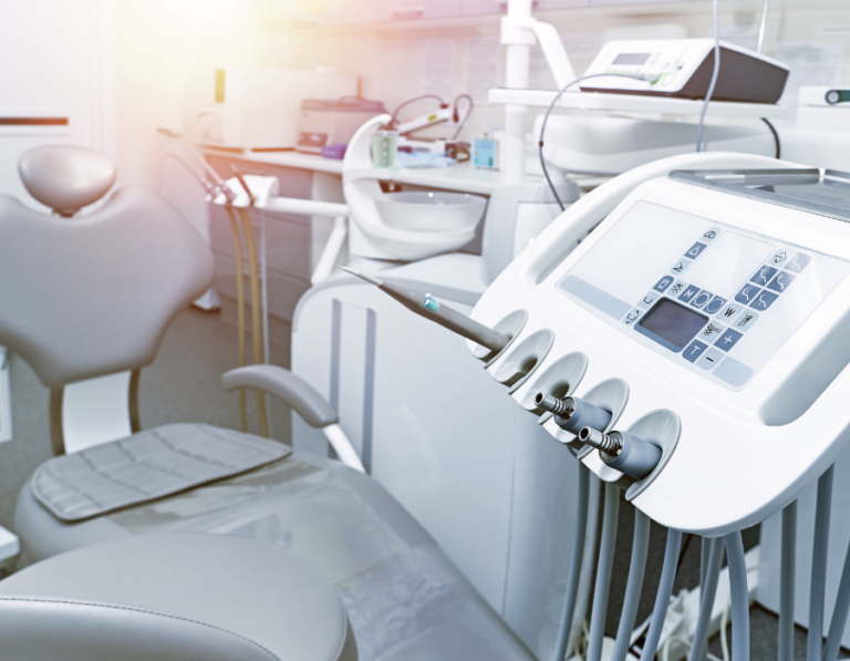 Brightly lit dental clinic showing a dental chair with attached tools and equipment console, featuring various handpieces and digital controls.