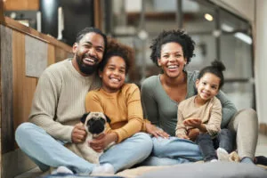 A happy family sitting together on the floor in their home, smiling at the camera. The family consists of a father, mother, two daughters, and a pug dog. They are casually dressed and appear joyful and relaxed in their living space.