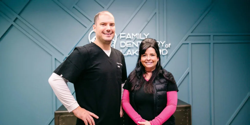 Two staff standing side by side in front of a wall with the text 'FAMILY DENTISTRY OF LAKEWOOD' visible in the background. One person is wearing black scrubs with a name tag, and the other is wearing a black vest over a pink long-sleeve shirt.